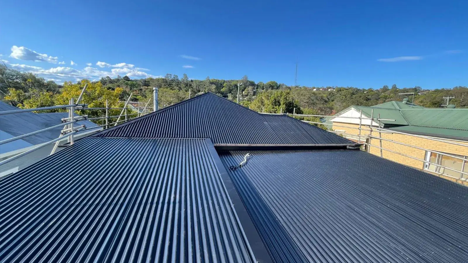 Newly installed dark corrugated metal roof with a central valley and scaffold edge protection on a hillside home