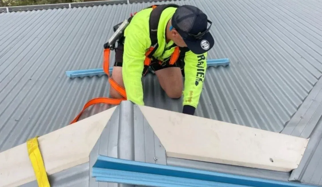 Roofer in hi-vis and a full safety harness kneeling on a light grey corrugated roof to fit a hip capping