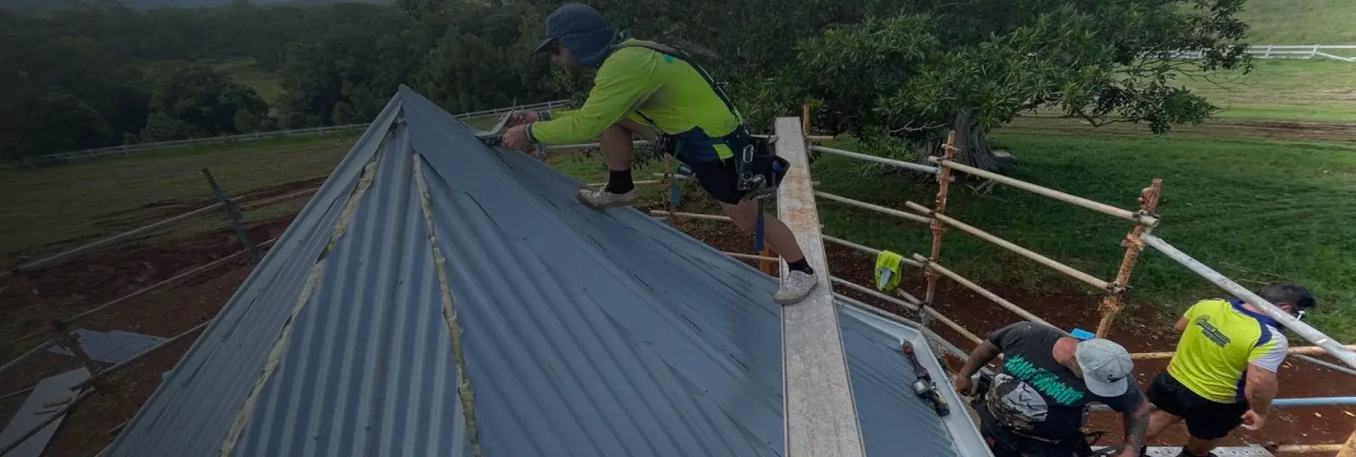 Roofing crew in hi-vis fixing dark grey corrugated sheets on a rural hip roof behind timber-rail edge protection