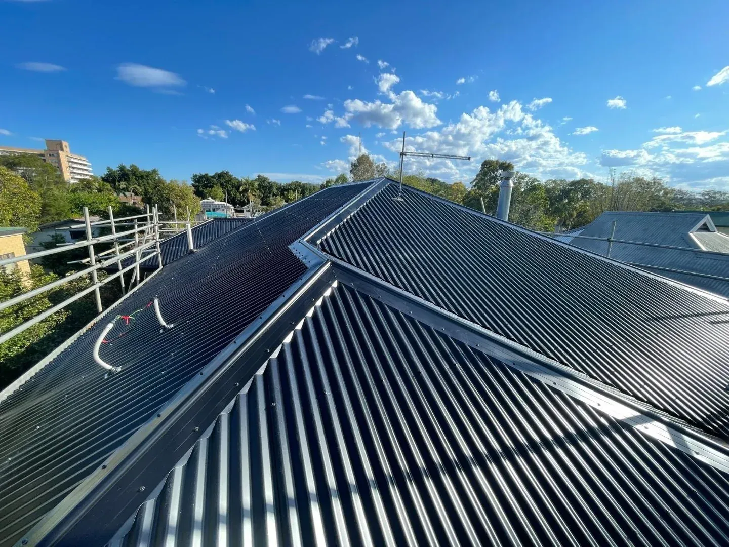 Dark corrugated metal re-roof with scaffold edge protection and a multi-storey building behind